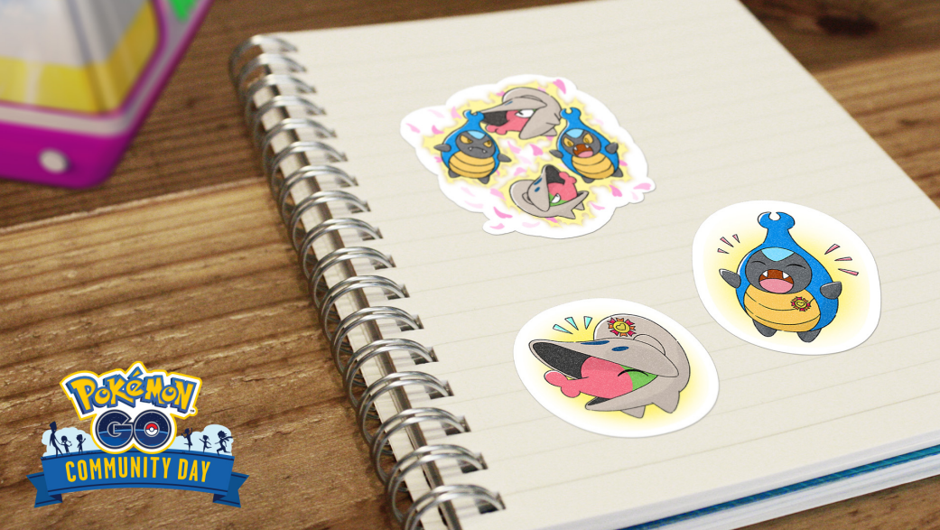 A notebook is shown on a desk with stickers featuring Karrablast and Shelmet stuck to it. In the top left corner is the edge of a Pokémon GO gift, and in the bottom left is the Pokémon GO Community Day logo